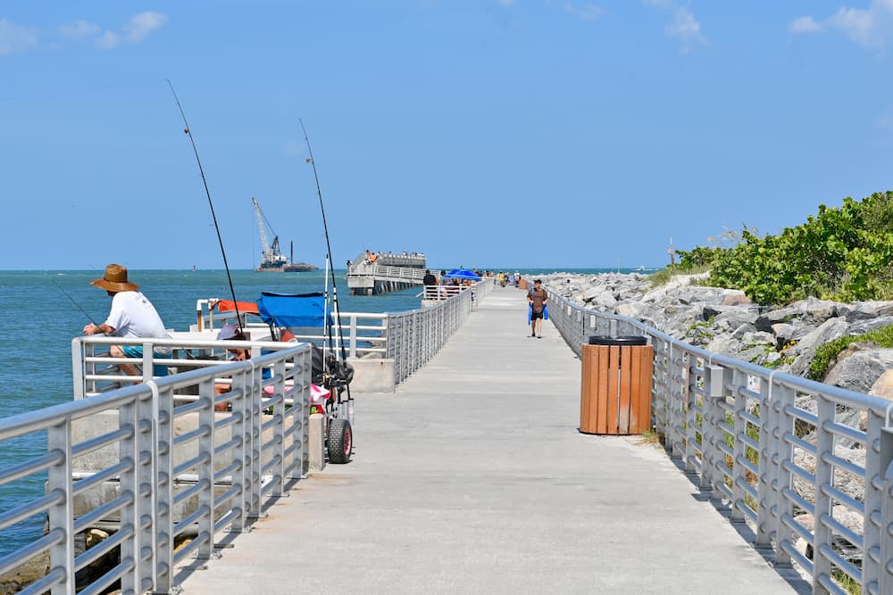 Anglers fishing along the railing of Jetty Park's fishing pier in Port Canaveral, with visitors walking the pier toward the water under a clear blue sky