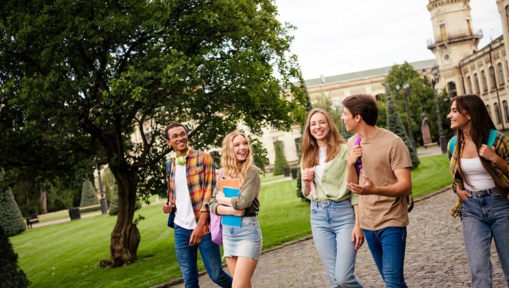 A group of five smiling high school students walking together on a college campus, carrying backpacks and notebooks near a tree-lined path.