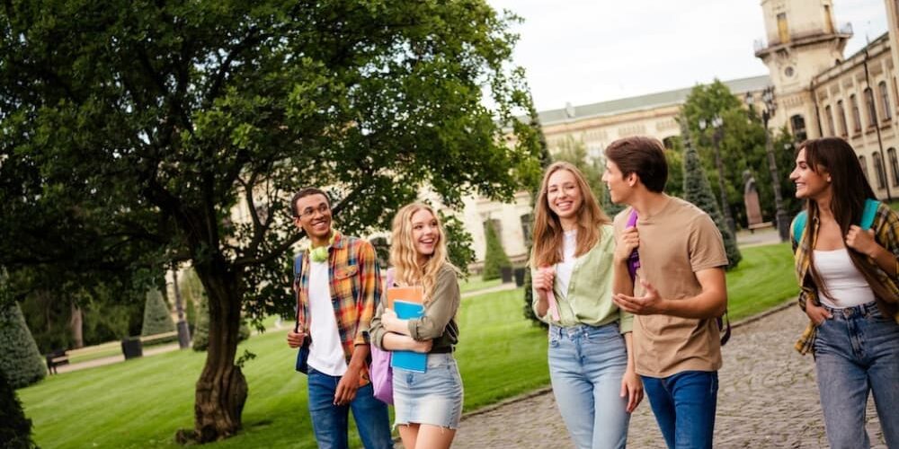 A group of five smiling high school students walking together on a college campus, carrying backpacks and notebooks near a tree-lined path.